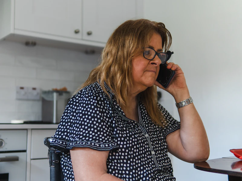 Mulher de cabelo claro e óculos falando ao telefone em uma cozinha clara