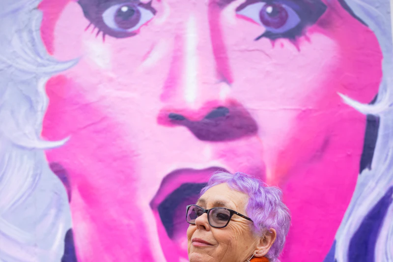 Mulher de cabelo lilás, óculos e casaco colorido, diante de um mural pintado